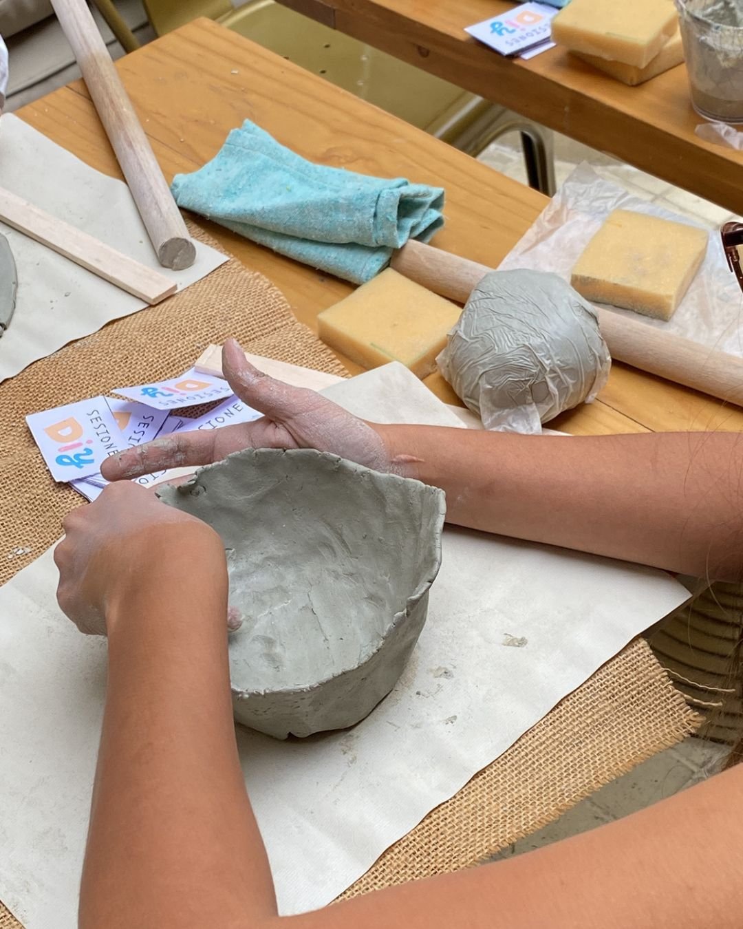 Manos dando forma a un cuenco de arcilla en uno de nuestros talleres de arte, con rodillo, esponjas y tarjetas Sesiones DIY