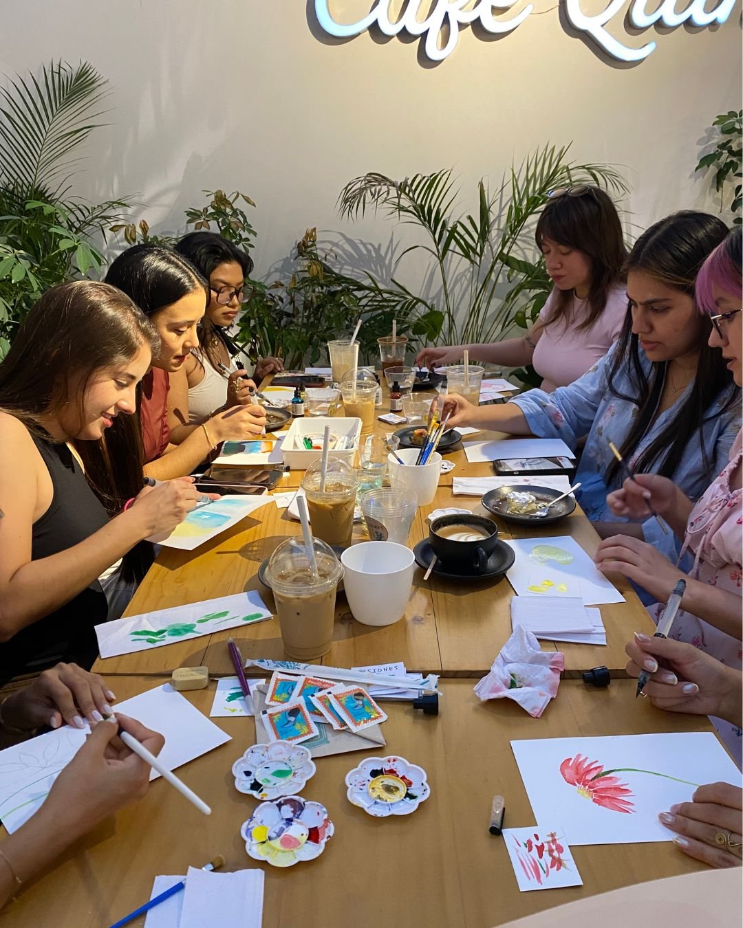 Grupo pintando con acuarelas en una mesa de madera con café helado y paletas de colores, en nuestros talleres de arte