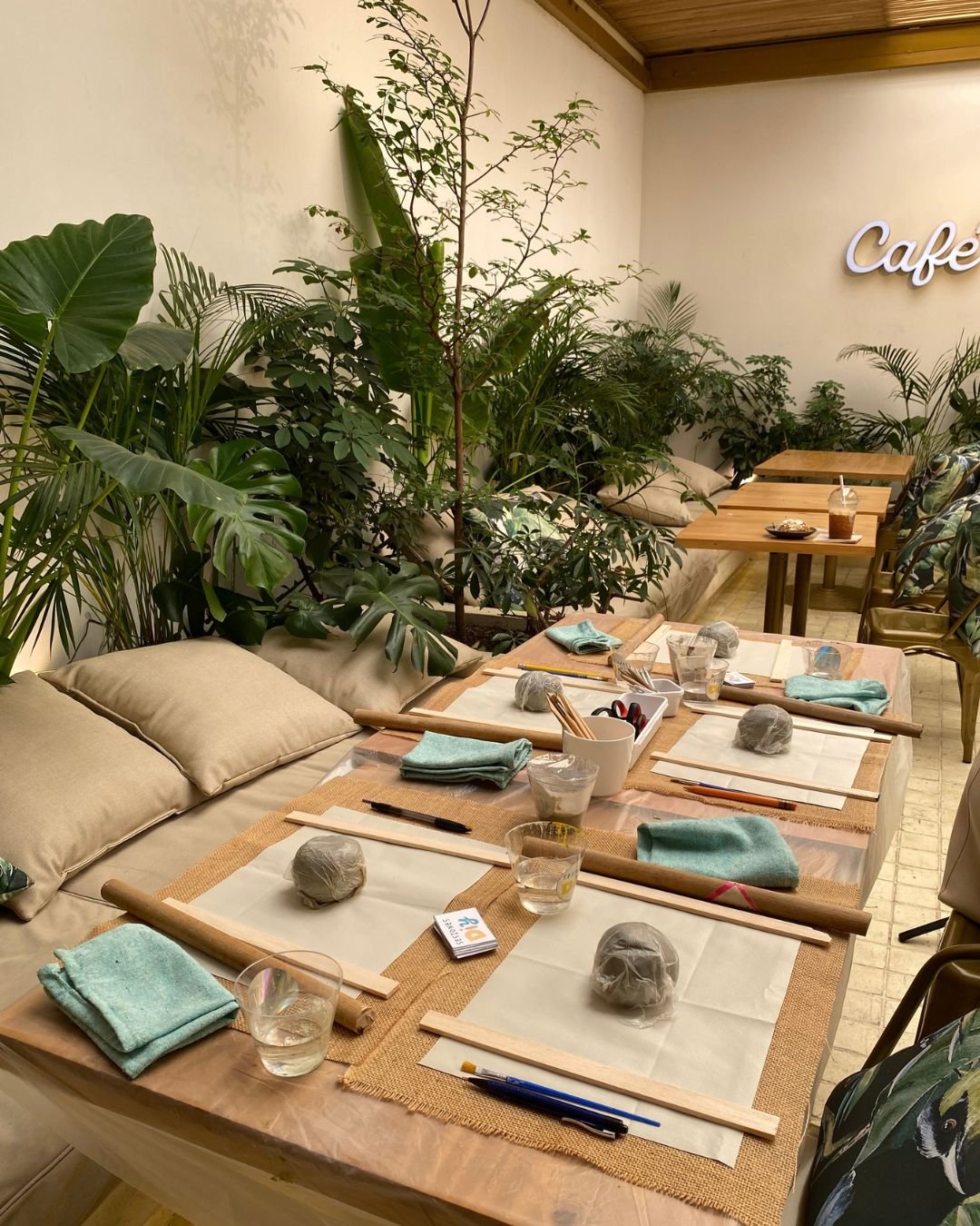 Mesa preparada con bolas de arcilla, rodillos y paños verdes en un café con plantas, lista para nuestros talleres de arte