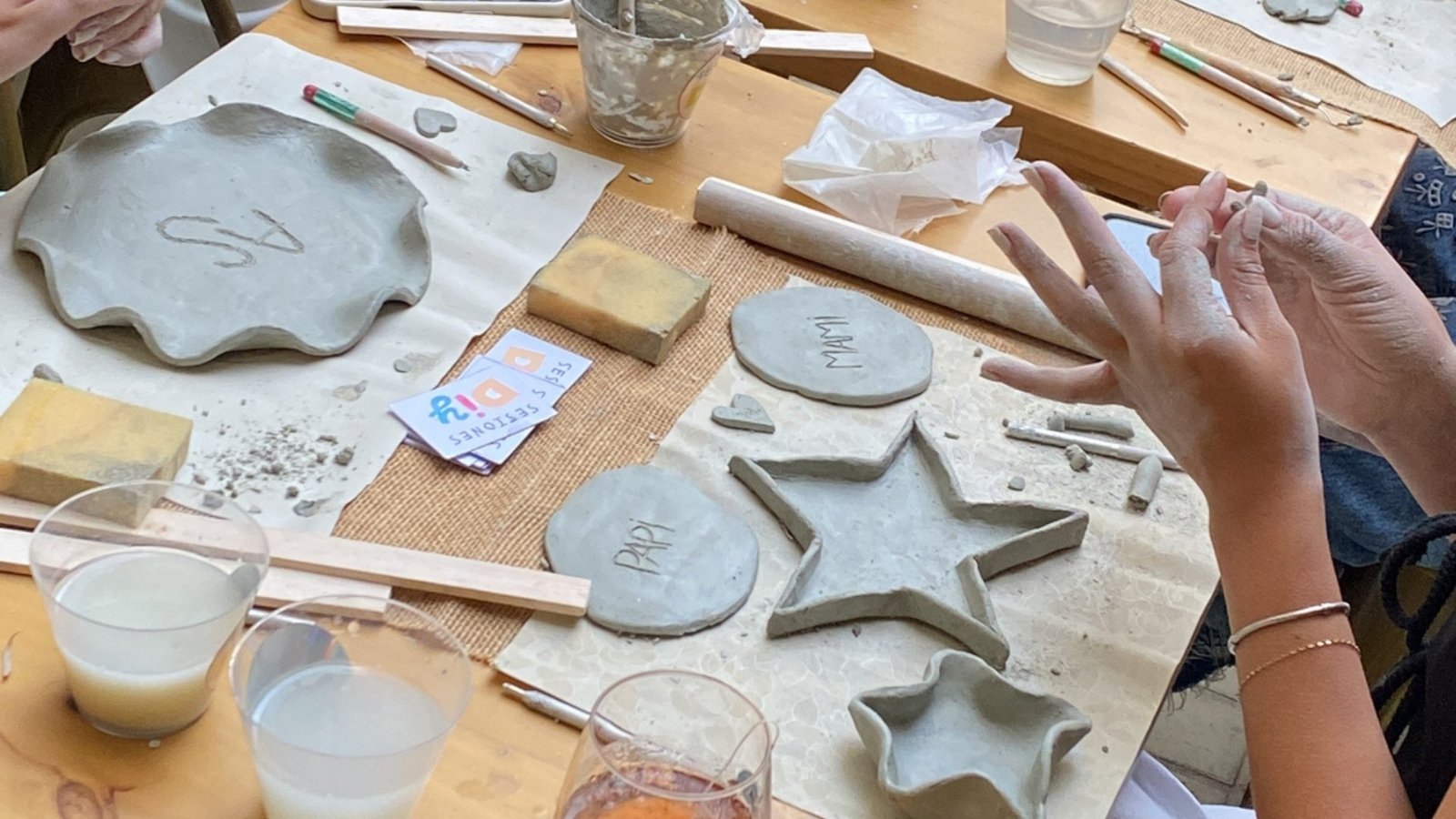 Manos moldeando arcilla en una mesa de nuestros talleres de arte en Medellín, con platos, una estrella y tarjetas Sesiones DIY